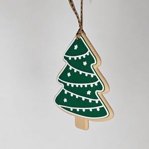 K&ouml;nnte beinhalten: Ein weihnachtsbaumf&ouml;rmiger Schmuck mit gr&uuml;nem Design und wei&szlig;en Akzenten. Der Schmuck ist wie ein Keks gestaltet, mit einem beigen Rand und einem braunen Band zum Aufh&auml;ngen. Der Baum hat wei&szlig;e Lichterketten und Sterndetails.