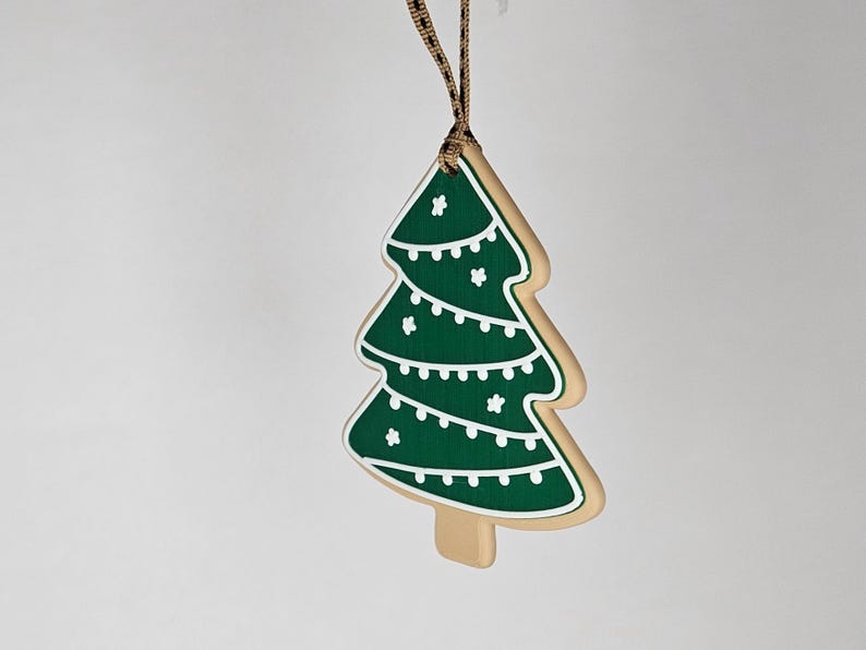K&ouml;nnte beinhalten: Ein weihnachtsbaumf&ouml;rmiger Schmuck mit gr&uuml;nem Design und wei&szlig;en Akzenten. Der Schmuck ist wie ein Keks gestaltet, mit einem beigen Rand und einem braunen Band zum Aufh&auml;ngen. Der Baum hat wei&szlig;e Lichterketten und Sterndetails.