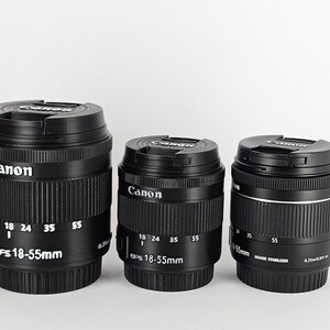 May include: Three black Canon EFS 18-55mm camera lenses. The lenses have a silver band around the middle and white markings indicating the focal length.