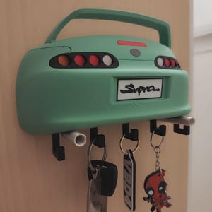 May include: A mint green car-shaped key holder with the word "Supra" on the back. The key holder has a handle and several black hooks holding keys and keychains. The keychains include a "Vibda" tag and a Deadpool figure.