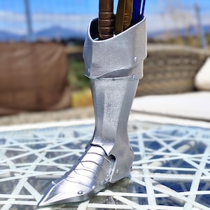 May include: A silver-coloured pen holder in the shape of a knight's boot, featuring a pointed toe and riveted details. It holds pens in brown, gold, and blue. The background is a blurred outdoor scene.