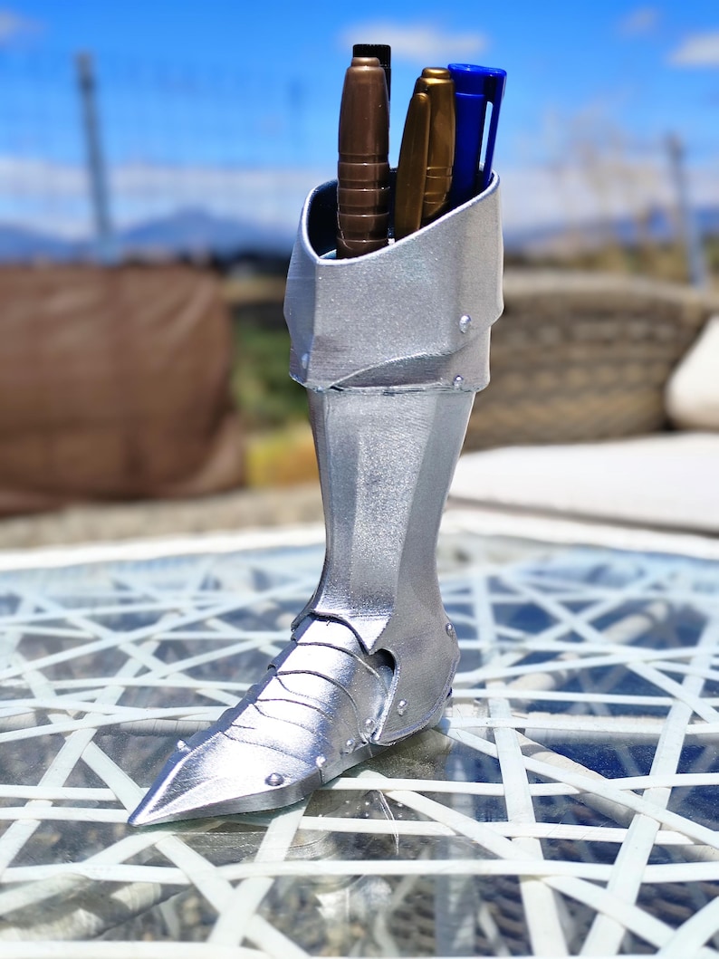 May include: A silver-coloured pen holder in the shape of a knight's boot, featuring a pointed toe and riveted details. It holds pens in brown, gold, and blue. The background is a blurred outdoor scene.