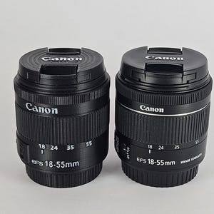 May include: Two black Canon EF-S 18-55mm camera lenses with lens caps. The lens on the left has a black finish, while the lens on the right has a silver band around the middle.