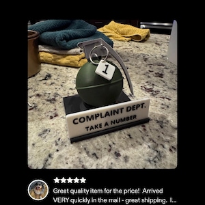 May include: A novelty item featuring a green grenade with a metal pull ring and a small tag with the number 1. Below it is a sign that reads "COMPLAINT DEPT. TAKE A NUMBER."