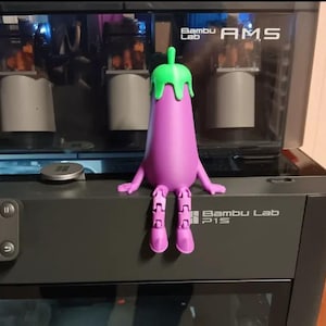 May include: A 3D-printed purple eggplant figurine with green dripping top, arms, and articulated legs. The figurine is sitting on a black 3D printer. The printer has the text "Bambu Lab P1S" on the front.