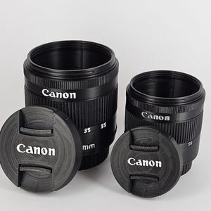 May include: Two black Canon camera lens caps with a black lens cap on each. The larger lens cap has the text "Canon" and "35 55 mm" printed on it. The smaller lens cap has the text "Canon" printed on it.