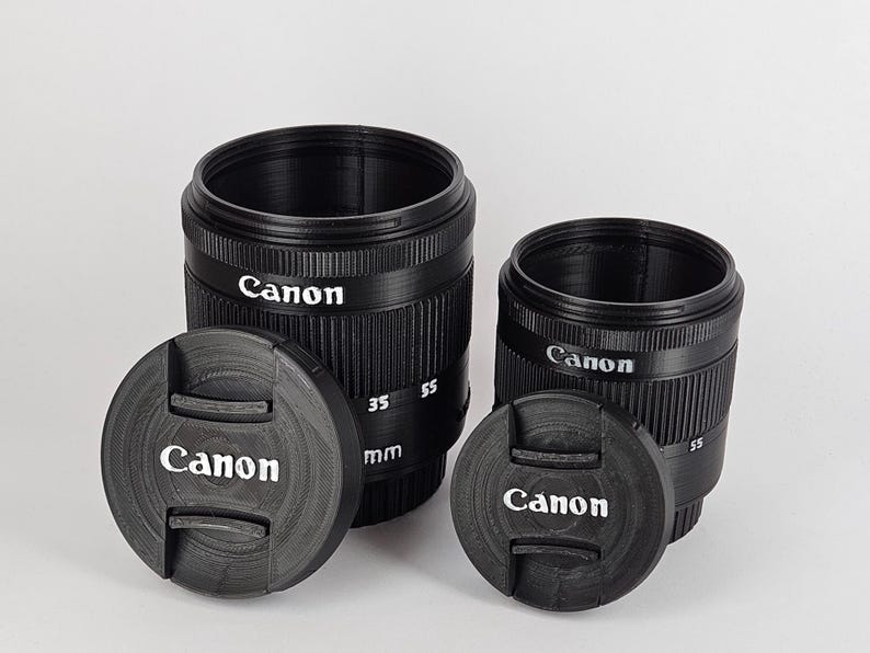 May include: Two black Canon camera lens caps with a black lens cap on each. The larger lens cap has the text "Canon" and "35 55 mm" printed on it. The smaller lens cap has the text "Canon" printed on it.
