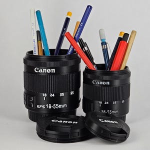 May include: Two black Canon camera lens containers with writing that says "Canon" and "EFS 18-55mm". The containers are filled with various colored pencils and pens.