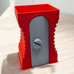 以下が含まれることがあります： 中央にネジが付いた、グレーの研磨刃を持つ赤いプラスチック製の鉛筆削り器。鉛筆削り器は側面にリブのデザインが施されています。