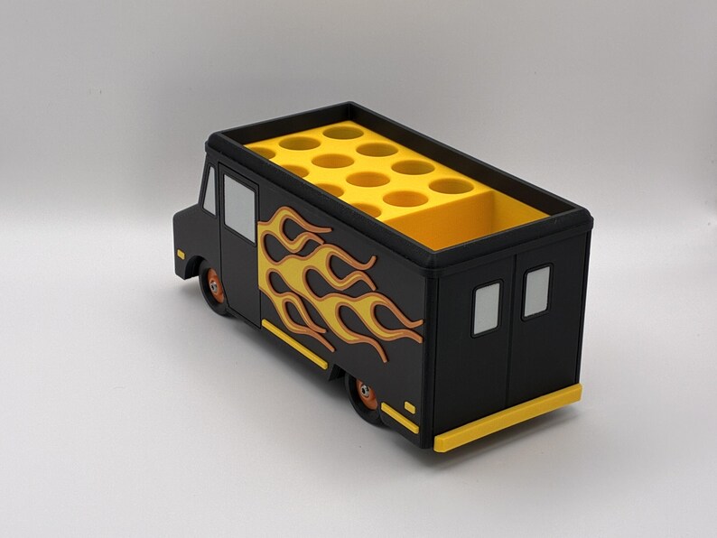 May include: A black and yellow food truck-shaped desk organizer. The organizer features a flame design on the side and a yellow interior with multiple circular compartments. The wheels and trim are also yellow.