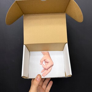 May include: An open cardboard box with a printed image of a hand making the "okay" gesture inside. The box is held by a person's hand against a black background. The box is brown with white interior walls.