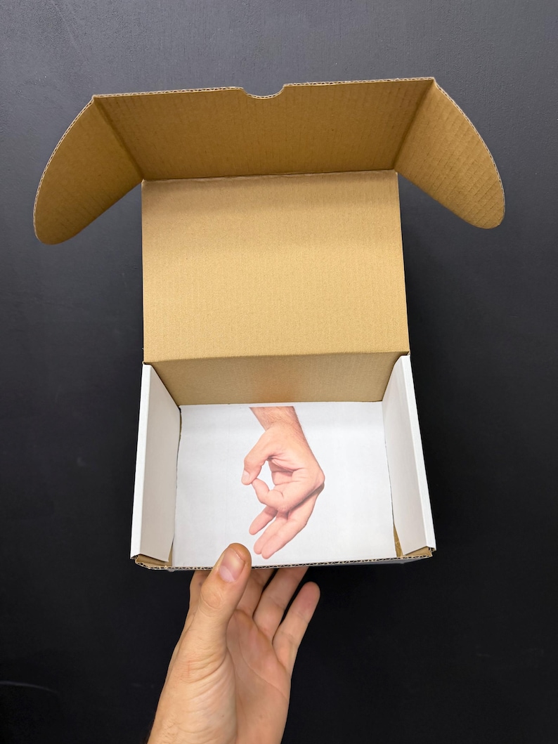 May include: An open cardboard box with a printed image of a hand making the "okay" gesture inside. The box is held by a person's hand against a black background. The box is brown with white interior walls.