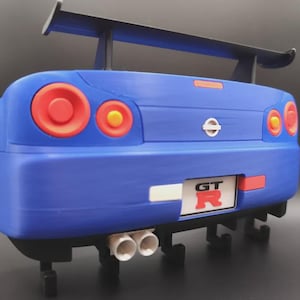 May include: A blue model car rear end with red taillights and a black spoiler. The car has a white license plate with the letters "GT" and the letter "R" in red. The car has two white exhaust pipes.