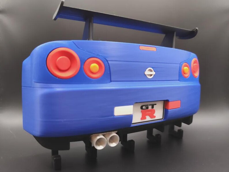 May include: A blue model car rear end with red taillights and a black spoiler. The car has a white license plate with the letters "GT" and the letter "R" in red. The car has two white exhaust pipes.