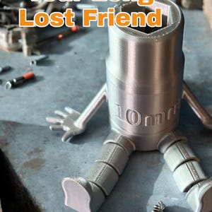May include: A silver 3D printed model of a 10mm socket wrench with legs and arms. The model is sitting on a textured surface. The text "Your Long Lost Friend" and "XL Version" are visible in the image.