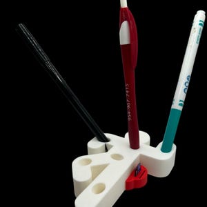 May include: White plastic pen holder shaped like a person with three holes for pens. A red pen, a black pen, and a green marker are in the holder. A red and white sticker is on the holder.