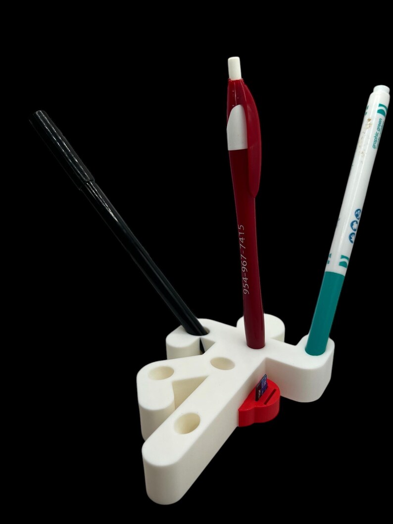 May include: White plastic pen holder shaped like a person with three holes for pens. A red pen, a black pen, and a green marker are in the holder. A red and white sticker is on the holder.