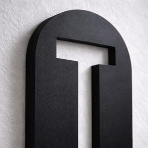 Op de afbeelding: Een matzwarte, abstracte wandkunst met een getextureerd oppervlak. Het ontwerp heeft een afgeronde bovenkant met een rechthoekige uitsparing en een verticaal rechthoekig element eronder, wat een moderne, geometrische esthetiek cre&euml;ert. Het kunstwerk is op een witte, getextureerde muur gemonteerd.
