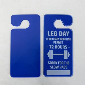 K&ouml;nnte beinhalten: Zwei blaue T&uuml;ranh&auml;nger. Einer ist leer, der andere tr&auml;gt den Text "LEG DAY TEMPORARY HOBBLING PERMIT - 72 HOURS -" mit einer Hantelgrafik und "SORRY FOR THE SLOW PACE" darunter.