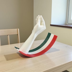 Op de afbeelding: Een witte sculptuur van een handgebaar, met de vingers tegen elkaar geknepen, wordt gecombineerd met een gebogen basis in de kleuren van de Italiaanse vlag: groen, wit en rood. De sculptuur wordt tentoongesteld op een lichtgekleurde houten tafel.