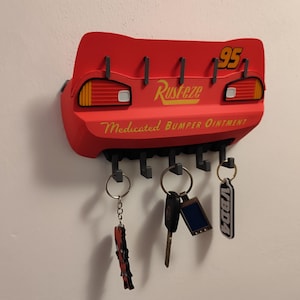 May include: A red key holder shaped like Lightning McQueen's rear end from the movie Cars. It features the number 95 and the word "Rusteze". The key holder has several hooks with keys attached.