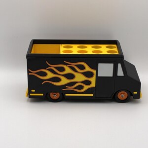 May include: A black food truck-shaped desk organizer with yellow and orange flame decals on the side. The top has multiple circular compartments and a separate rectangular section. The wheels are orange.