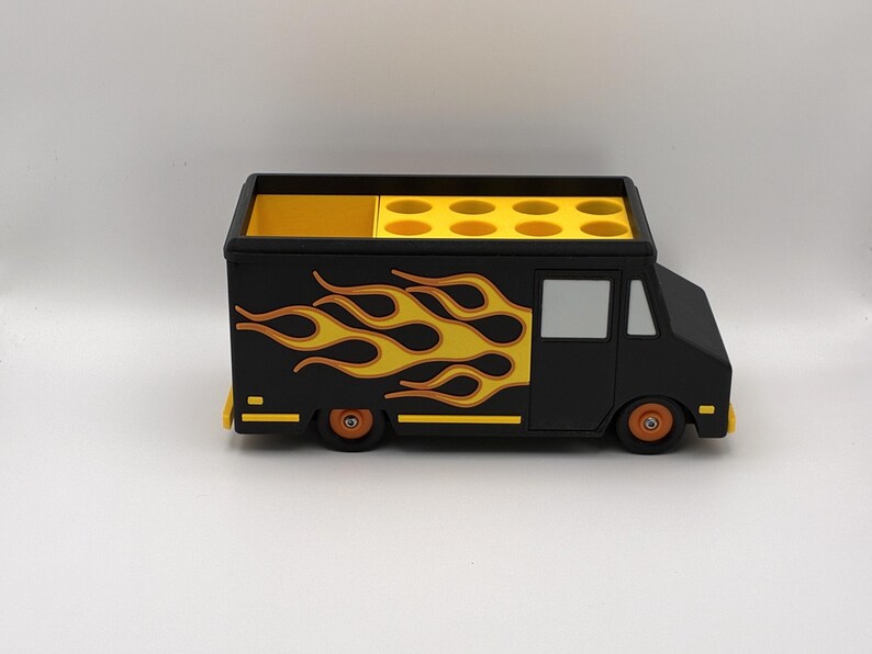 May include: A black food truck-shaped desk organizer with yellow and orange flame decals on the side. The top has multiple circular compartments and a separate rectangular section. The wheels are orange.