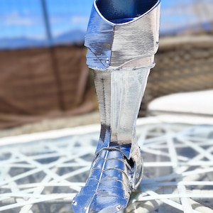 May include: A silver decorative boot, possibly a sculpture or figurine. The boot has a metallic sheen and is detailed with rivets and segmented plates, suggesting armour or a historical design. The boot is displayed outdoors.