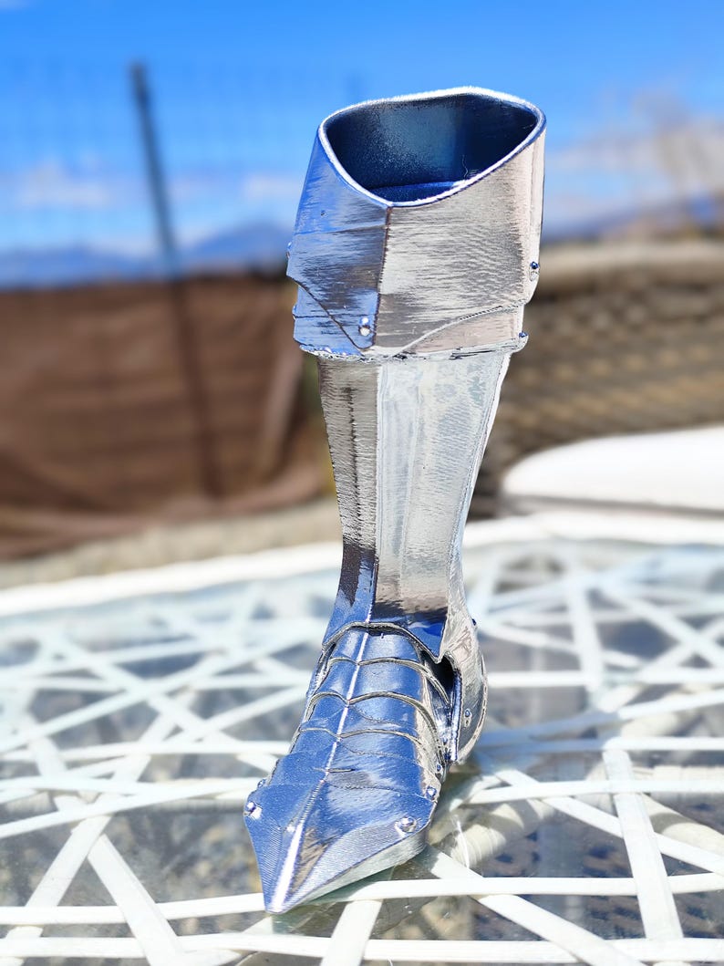 May include: A silver decorative boot, possibly a sculpture or figurine. The boot has a metallic sheen and is detailed with rivets and segmented plates, suggesting armour or a historical design. The boot is displayed outdoors.