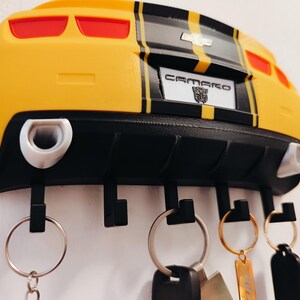 Puede incluir: Llavero Camaro amarillo y negro con una placa que dice "CAMARO". El llavero tiene cuatro ganchos negros y llaveros plateados con llaves adjuntas. El llavero est&aacute; montado en una pared blanca.