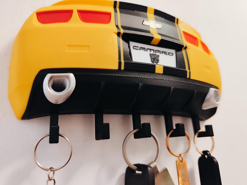 Puede incluir: Llavero Camaro amarillo y negro con una placa que dice "CAMARO". El llavero tiene cuatro ganchos negros y llaveros plateados con llaves adjuntas. El llavero est&aacute; montado en una pared blanca.