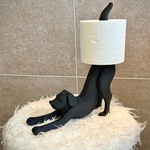 Pode incluir: Um porta-papel higi&ecirc;nico em forma de cachorro preto. O cachorro est&aacute; em uma pose brincalhona, segurando um rolo de papel higi&ecirc;nico branco. O suporte est&aacute; em uma superf&iacute;cie branca e fofa, com um fundo de azulejos de tons neutros.