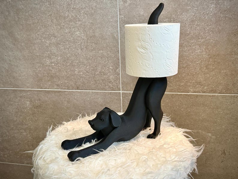 Pode incluir: Um porta-papel higi&ecirc;nico em forma de cachorro preto. O cachorro est&aacute; em uma pose brincalhona, segurando um rolo de papel higi&ecirc;nico branco. O suporte est&aacute; em uma superf&iacute;cie branca e fofa, com um fundo de azulejos de tons neutros.