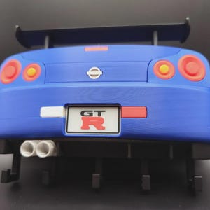 May include: A blue toy car with a rear spoiler, red and yellow taillights, and dual exhaust pipes. The car has a license plate that reads "GT" in red letters. The Nissan logo is visible.