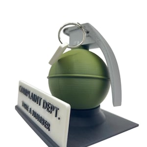 May include: A novelty desk accessory featuring a green grenade with a gray handle and ring, resting on a black base. A white sign with the text "COMPLAINT DEPT. TAKE A NUMBER" is positioned in front of the grenade.