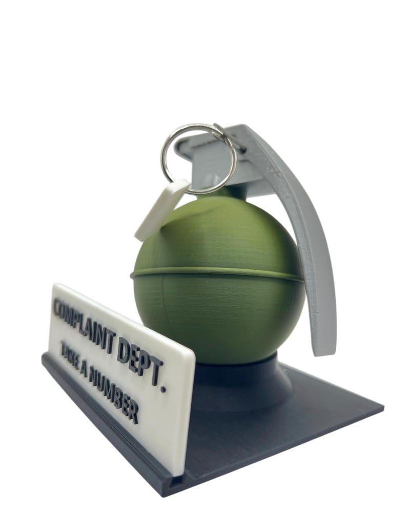May include: A novelty desk accessory featuring a green grenade with a gray handle and ring, resting on a black base. A white sign with the text "COMPLAINT DEPT. TAKE A NUMBER" is positioned in front of the grenade.