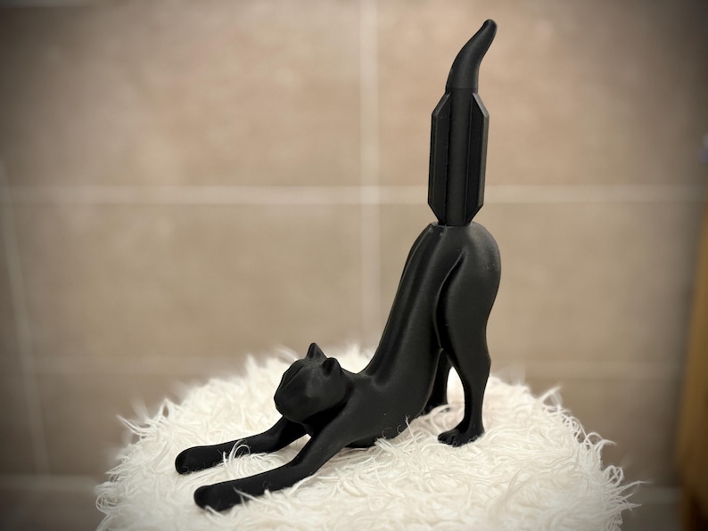 Pode incluir: Uma estatueta de gato preto em pose de alongamento. O gato est&aacute; de quatro, com as costas arqueadas e a cauda levantada. A estatueta &eacute; feita de um material preto s&oacute;lido e assenta numa superf&iacute;cie branca e fofa.