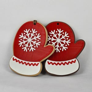 May include: Two red mitten-shaped Christmas ornaments with white snowflake designs. Each mitten has a white cuff with a red zigzag pattern. The ornaments are made of a textured material and have a hole at the top for hanging.
