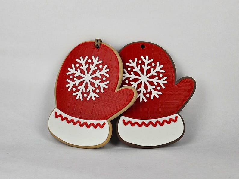 May include: Two red mitten-shaped Christmas ornaments with white snowflake designs. Each mitten has a white cuff with a red zigzag pattern. The ornaments are made of a textured material and have a hole at the top for hanging.