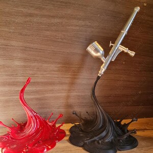 May include: A red splash sculpture and a black splash sculpture supporting a silver airbrush. The red splash is on the left, and the black splash is on the right. The airbrush is angled towards the right.