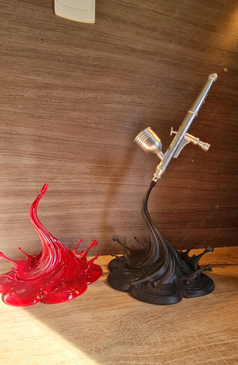 May include: A red splash sculpture and a black splash sculpture supporting a silver airbrush. The red splash is on the left, and the black splash is on the right. The airbrush is angled towards the right.