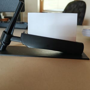 May include: A black business card holder with a rectangular base and a curved top. The holder is designed to hold a white business card. The holder is on a light brown surface.
