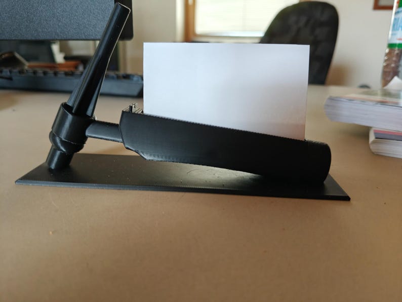 May include: A black business card holder with a rectangular base and a curved top. The holder is designed to hold a white business card. The holder is on a light brown surface.