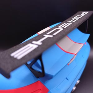 K&ouml;nnte beinhalten: Ein blaues und rotes Modellauto mit einem schwarzen Spoiler. Der Spoiler tr&auml;gt das Wort "PORSCHE" in wei&szlig;en Buchstaben. Das Auto hat einen grauen Streifen und rote Akzente.