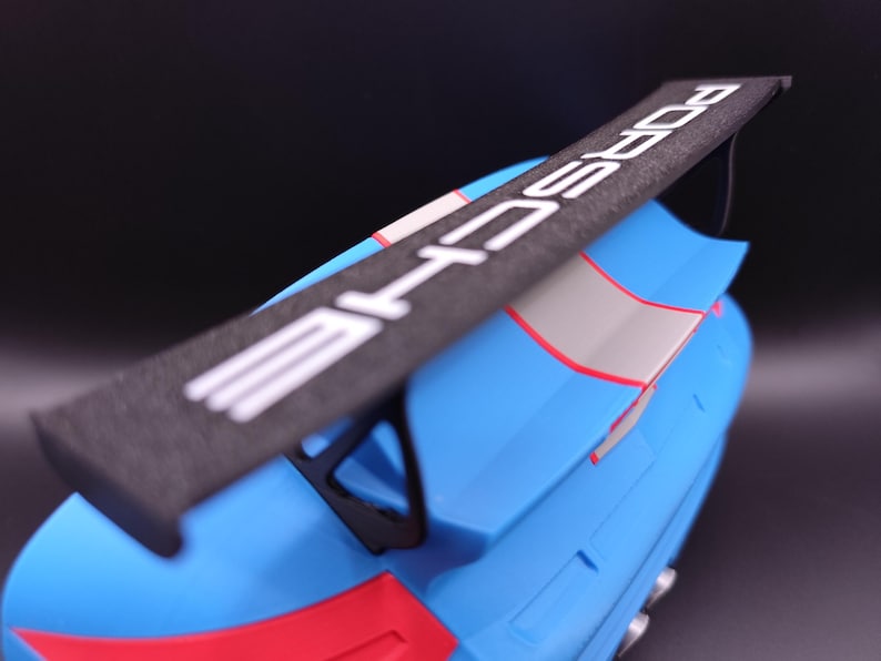 K&ouml;nnte beinhalten: Ein blaues und rotes Modellauto mit einem schwarzen Spoiler. Der Spoiler tr&auml;gt das Wort "PORSCHE" in wei&szlig;en Buchstaben. Das Auto hat einen grauen Streifen und rote Akzente.
