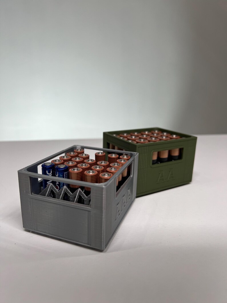 May include: Two 3D-printed battery organisers, one grey and one olive green, each filled with AA batteries. The grey organiser has a visible "AAA" label. The batteries are arranged neatly within the containers, showcasing a practical storage solution.