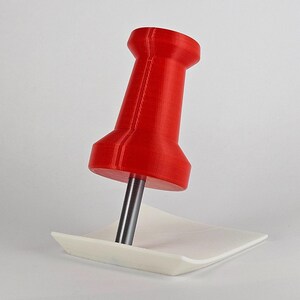 May include: A large, red push pin with a silver pin is inserted into a small, white, square paper. The push pin is a vibrant red color and has a classic design. The paper is plain and provides a contrasting background. Office supplies.