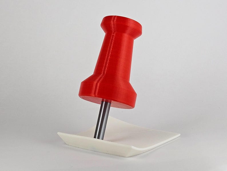 May include: A large, red push pin with a silver pin is inserted into a small, white, square paper. The push pin is a vibrant red color and has a classic design. The paper is plain and provides a contrasting background. Office supplies.