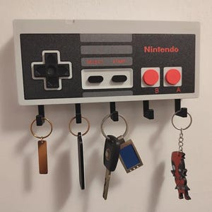 May include: A wall-mounted key holder designed as a Nintendo controller. The key holder is grey, black, and red, with the Nintendo logo. It has five black hooks for keys, with various keyrings attached.