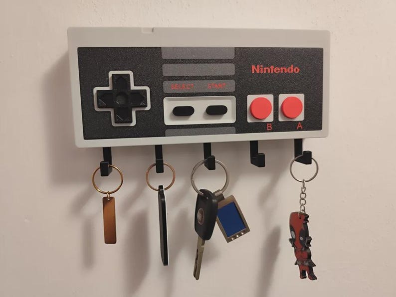 May include: A wall-mounted key holder designed as a Nintendo controller. The key holder is grey, black, and red, with the Nintendo logo. It has five black hooks for keys, with various keyrings attached.